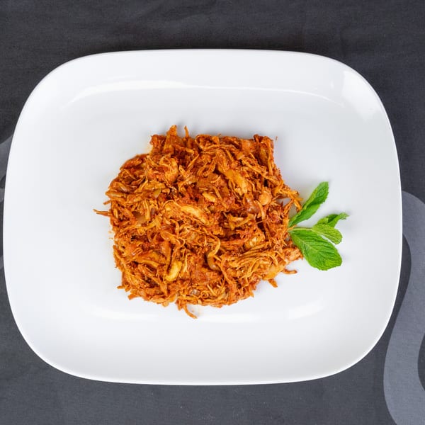 Shredded chicken in a rich red sauce with a sprig of mint on a white rectangular plate.