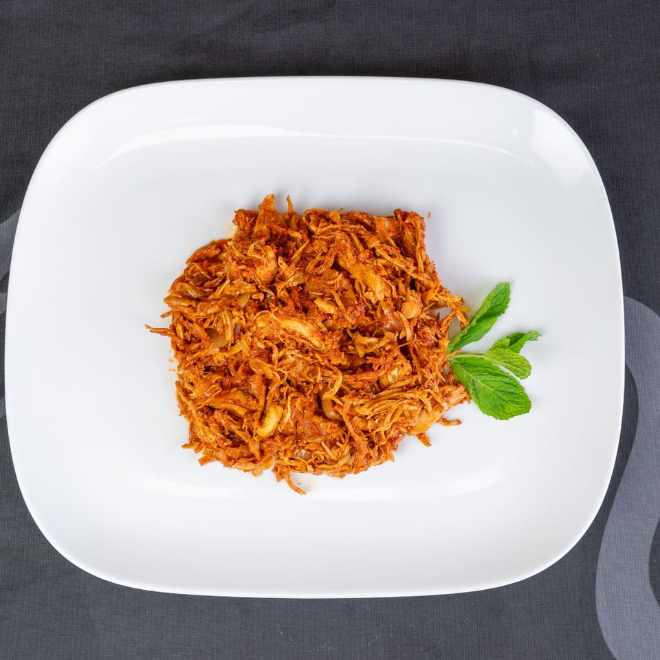 Shredded chicken in a rich red sauce with a sprig of mint on a white rectangular plate.