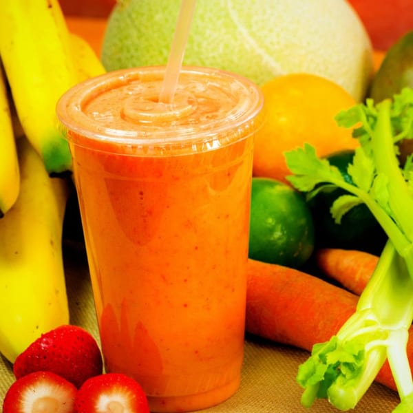 Orange smoothie with a straw, surrounded by fresh fruits and vegetables like bananas, strawberries, and carrots.