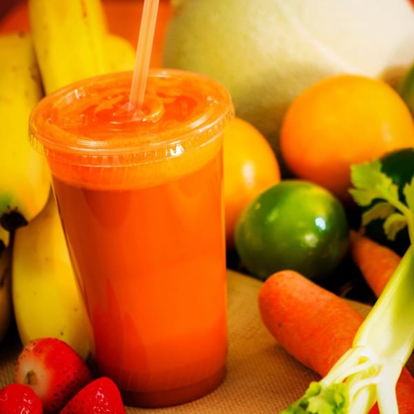 Freshly squeezed orange juice in a clear cup with a straw, surrounded by fruits and vegetables.