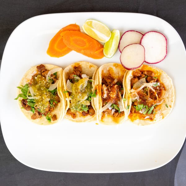 Four street tacos with meat, salsa, onions, cilantro, and sides of carrots, limes, and radishes.
