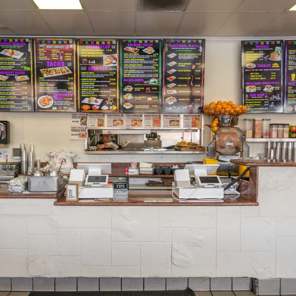 A bustling Mexican restaurant counter with menu boards, an orange juicer, and a chef preparing food.