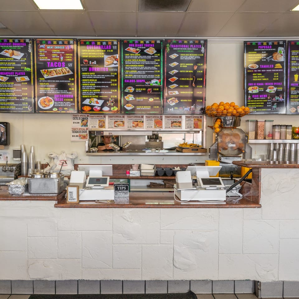 A bustling Mexican restaurant counter with menu boards, an orange juicer, and a chef preparing food.
