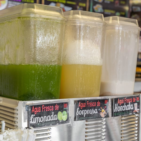 Three clear beverage dispensers filled with green, yellow, and white agua frescas, labeled Limonada, Sorpresa, and Horchata.