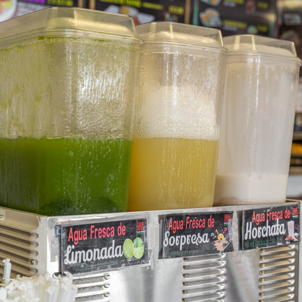 Three clear beverage dispensers filled with green, yellow, and white agua frescas, labeled Limonada, Sorpresa, and Horchata.