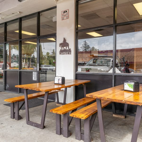 Two empty wooden picnic tables with benches sit outside a restaurant with large windows.