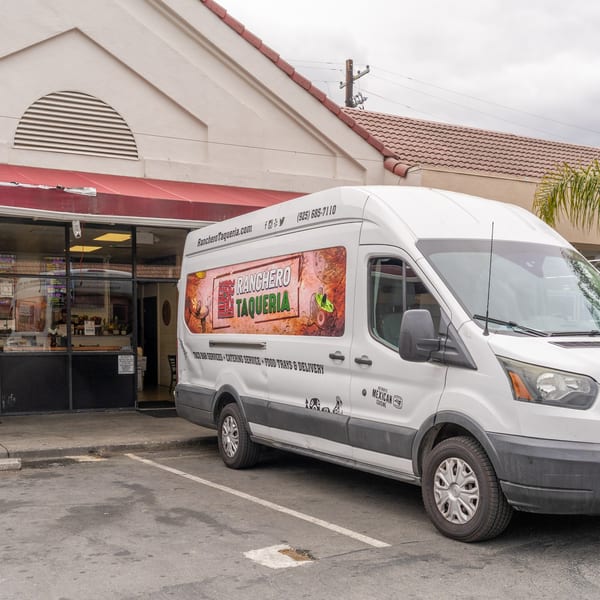 A white Ranchero Taqueria delivery van parked outside the restaurant with a red awning.