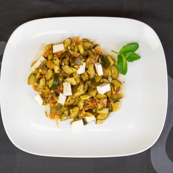 A white plate holds a serving of cooked zucchini and tomatoes with feta cheese and a mint garnish.