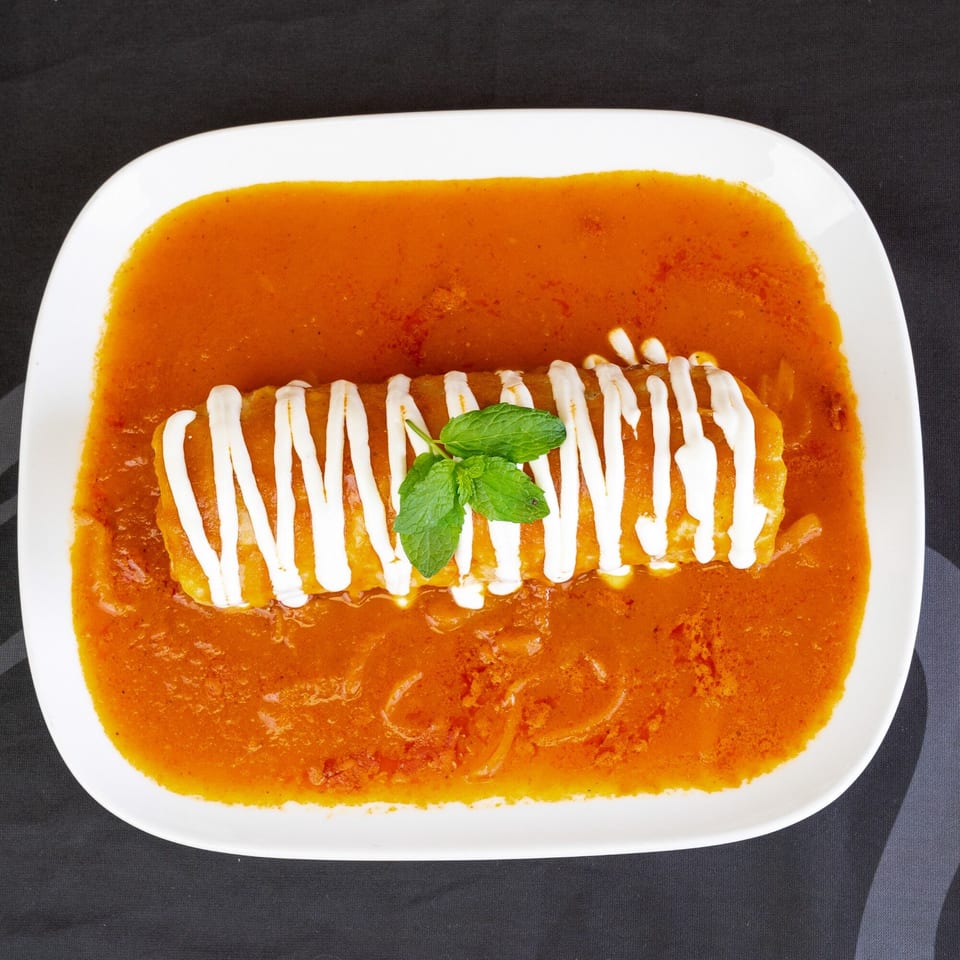 A white rectangular plate holds a burrito covered in red sauce, sour cream, and mint leaves.