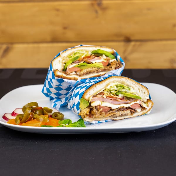 Two large torta sandwiches with various fillings, wrapped in blue and white checkered paper, on a white plate with pickled vegetables.