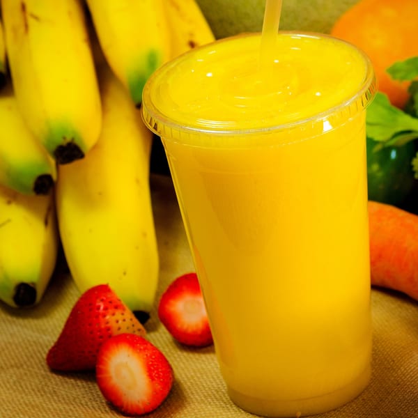 Bright yellow smoothie in a clear cup with a straw, surrounded by fresh bananas and strawberries.