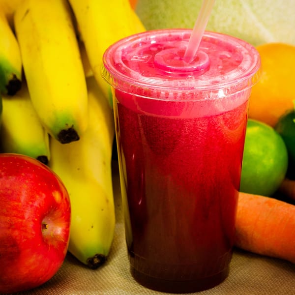 Bright red juice in a clear plastic cup with a straw, surrounded by fresh fruits and vegetables.