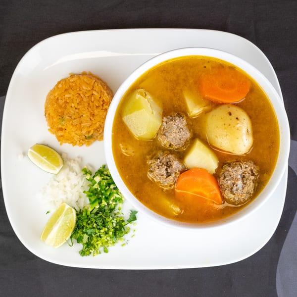 Albondigas soup with meatballs, carrots, and potatoes, served with rice, lime, and chopped onions.