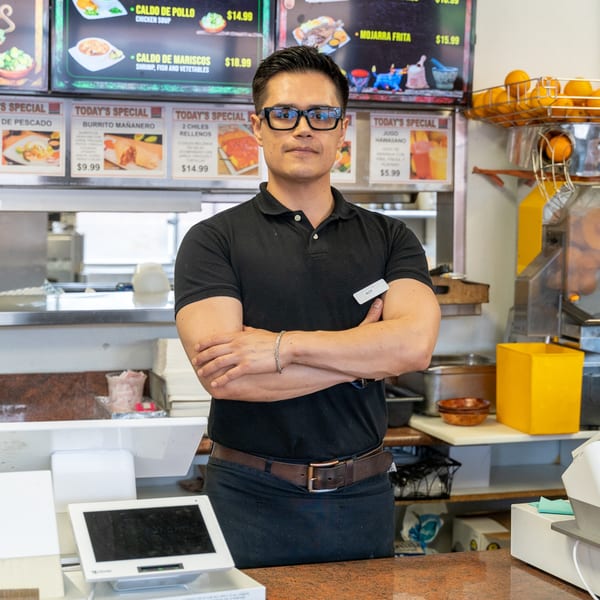 A man in a black polo shirt and apron stands with crossed arms behind a restaurant counter.