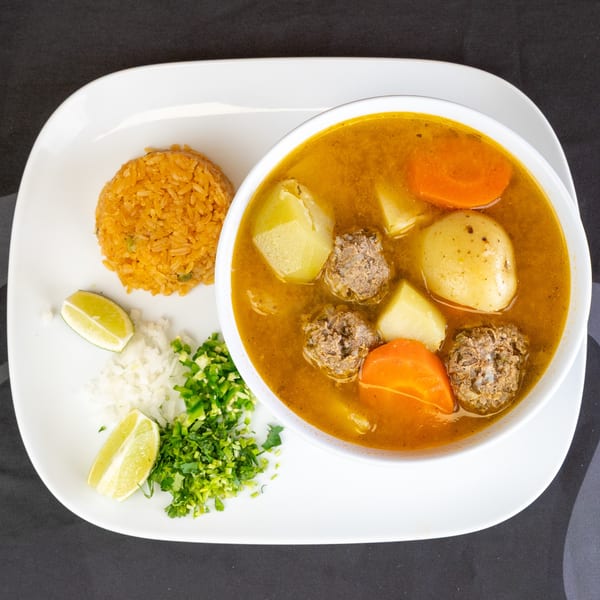Albondigas soup with meatballs, vegetables, and a side of rice, lime, and chopped herbs.