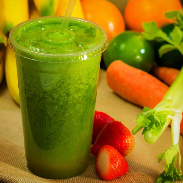 Green smoothie in a plastic cup with a straw, surrounded by fresh fruits and vegetables.