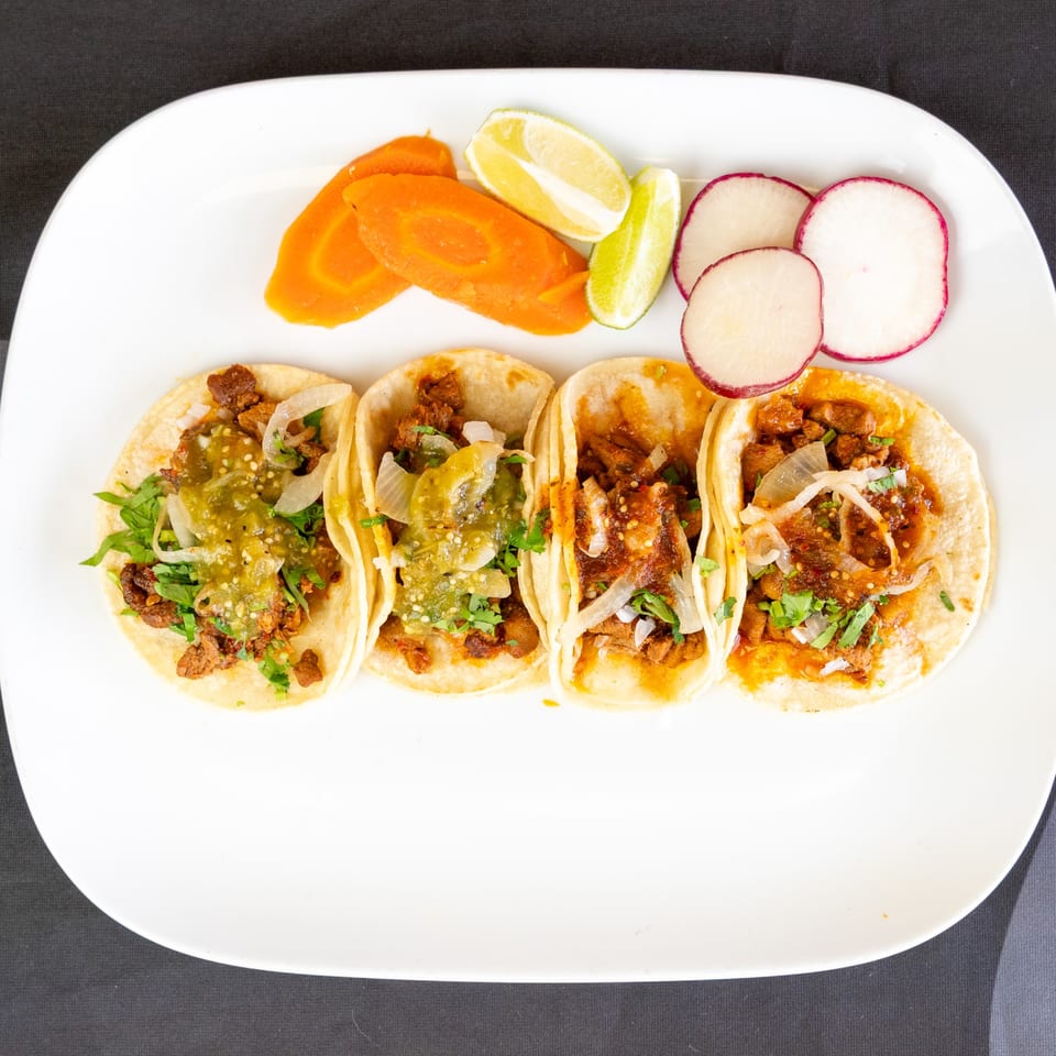 Four street tacos with meat, onions, cilantro, and salsa, served with lime, carrots, and radishes.